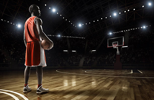 Basketball+player+standing+holding+basketball+on+an+indoor+floodlit+basketball+court+full+of+spectators.